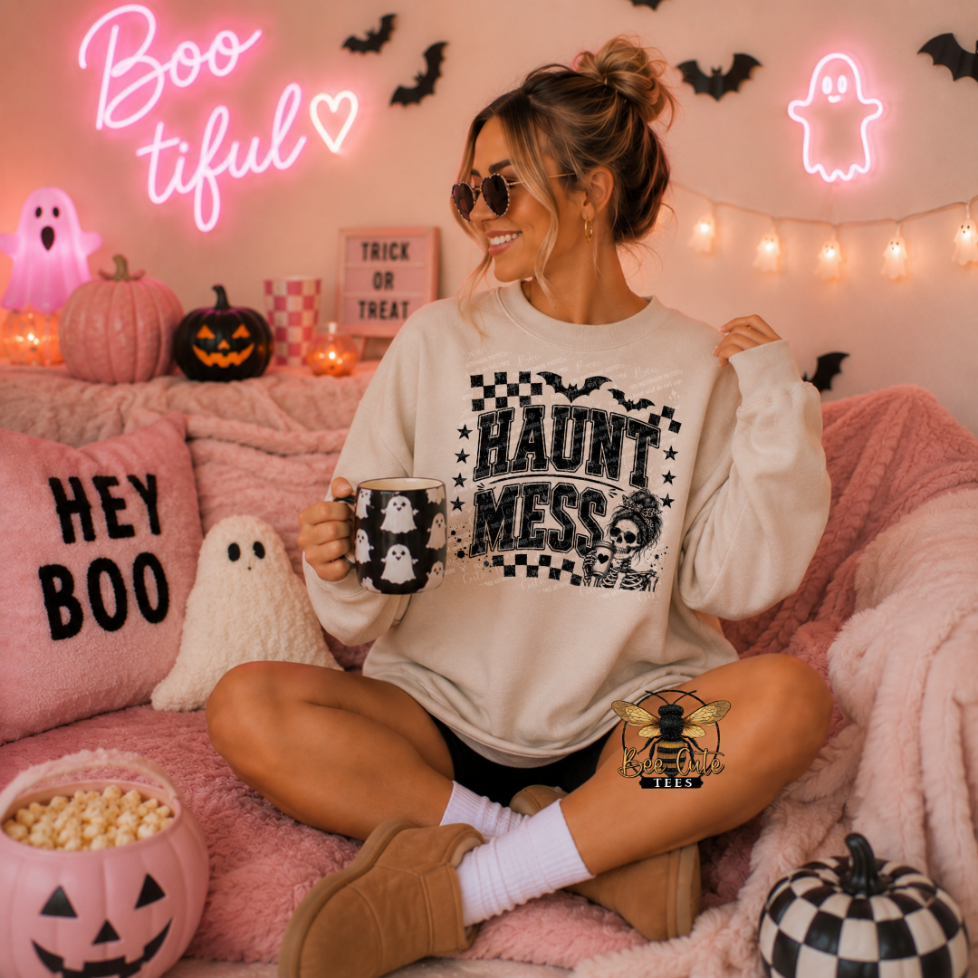A woman sits on a pink couch in a cozy Halloween-themed room, wearing a cream sweatshirt with a black 'HAUNT MESS' graphic and holding a mug. The room is decorated with pink neon signs, string lights, pumpkins, and a 'HEY BOO' pillow.