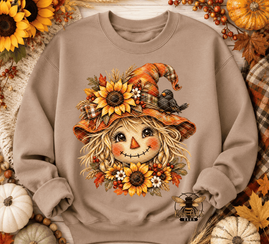 A beige crewneck sweatshirt featuring a detailed autumn scarecrow graphic with sunflowers, a plaid hat, a small bird, and fall leaves. The sweatshirt is styled in a flat-lay with seasonal props like pumpkins, sunflowers, and plaid fabric.