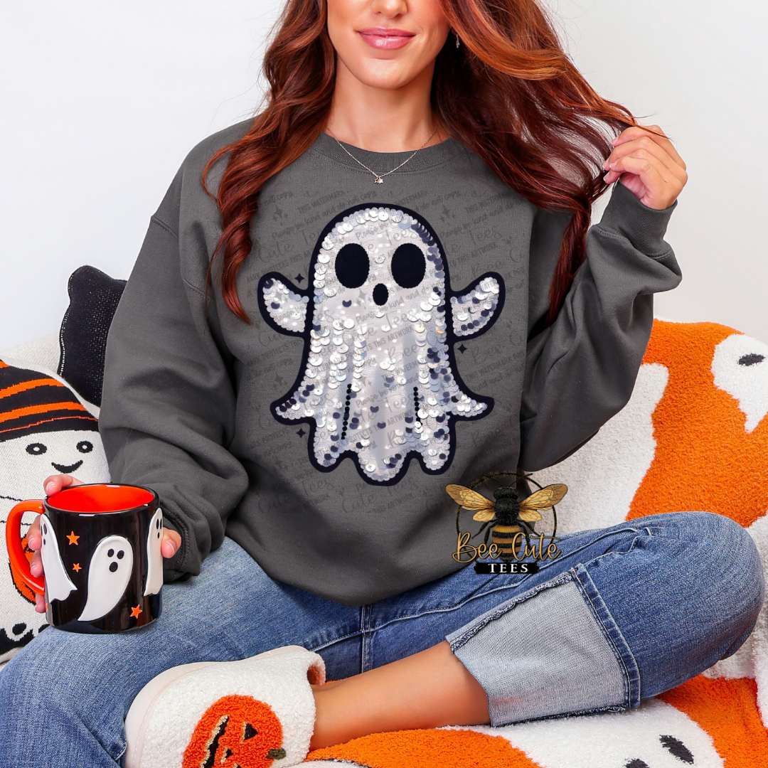 A woman wearing a dark grey crewneck sweatshirt with a large black and gray sequined ghost graphic on the front, paired with blue jeans and a black mug with a white ghost design. She is sitting on an orange and white blanket with Halloween-themed pillows.