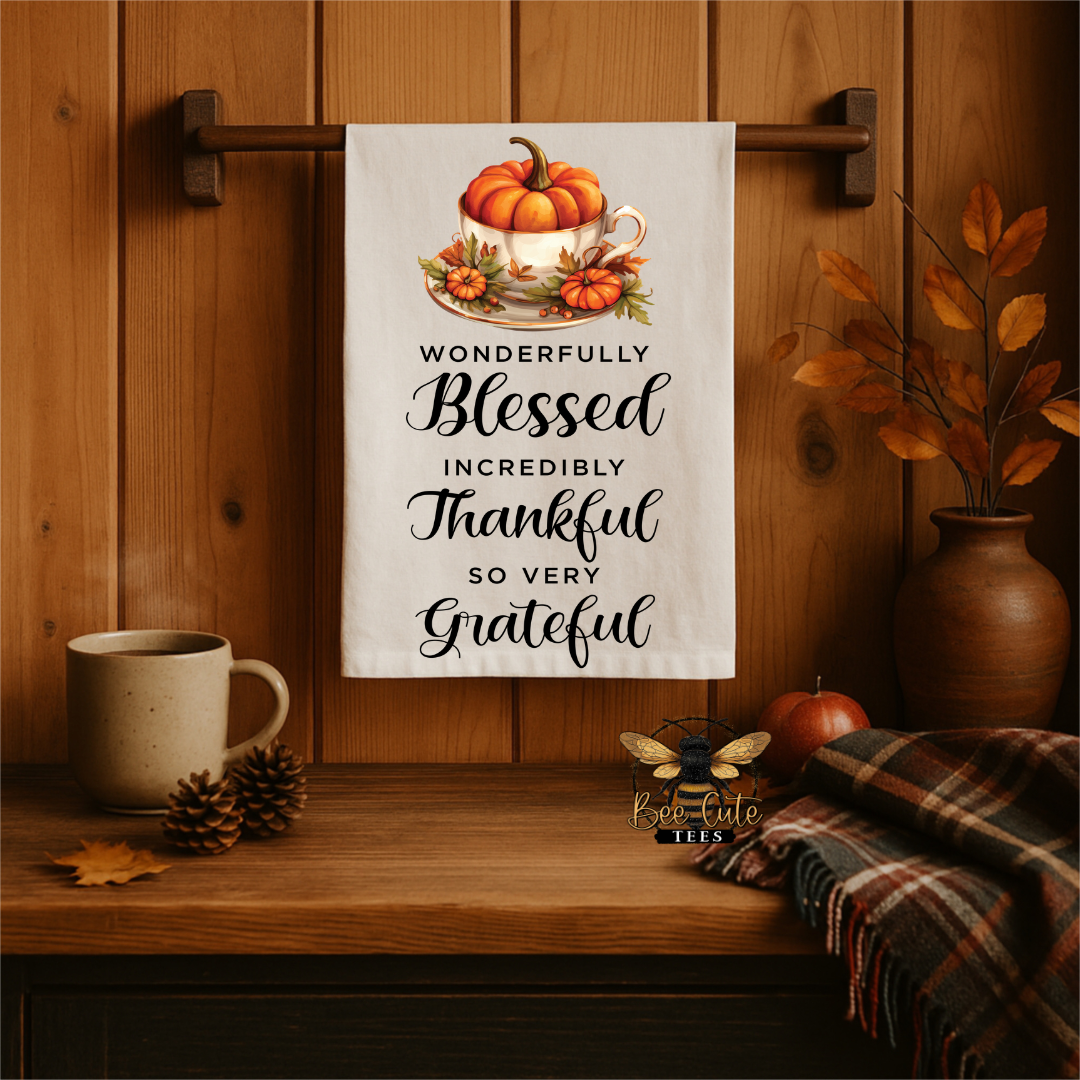 A white kitchen towel with a fall-themed illustration of pumpkins in a teacup and the text 'WONDERFULLY Blessed INCREDIBLY Thankful SO VERY Grateful' hanging on a wooden wall rack in a cozy autumn kitchen setting.