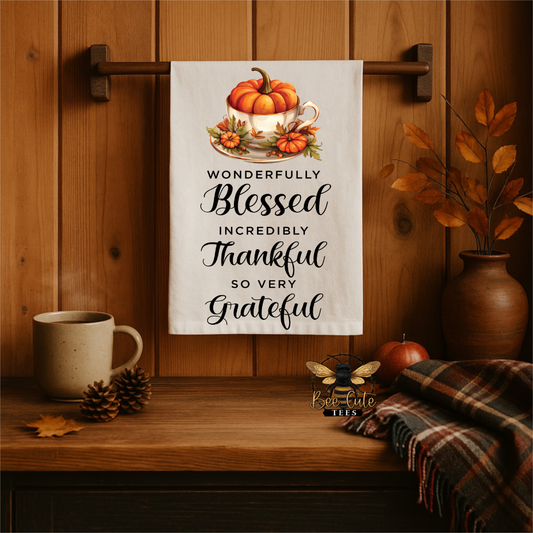 A white kitchen towel with a fall-themed illustration of pumpkins in a teacup and the text 'WONDERFULLY Blessed INCREDIBLY Thankful SO VERY Grateful' hanging on a wooden wall rack in a cozy autumn kitchen setting.