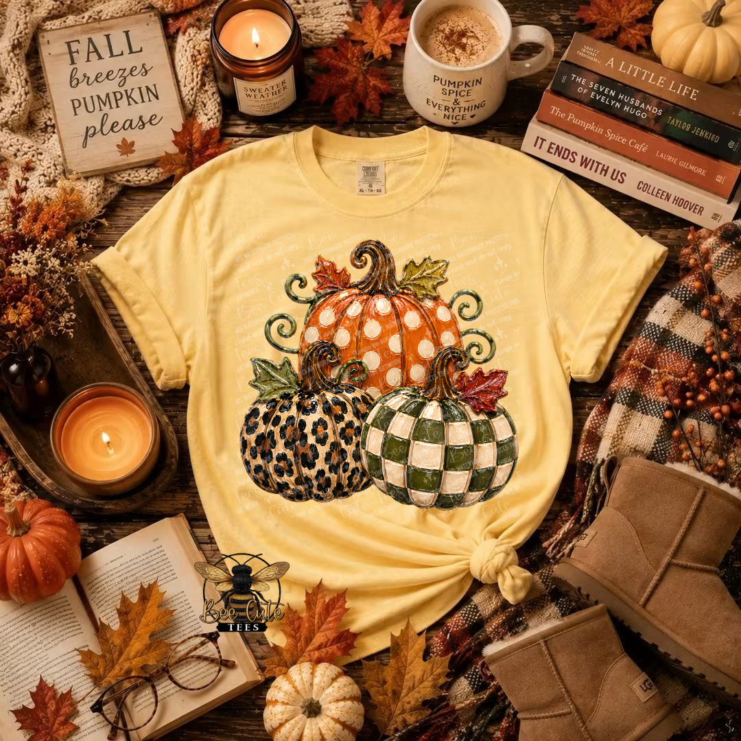 A yellow graphic t-shirt featuring three decorative pumpkins (orange polka dot, leopard print, and green-and-white checkered), styled in a cozy autumn flat-lay with candles, fall leaves, books, and a mug.