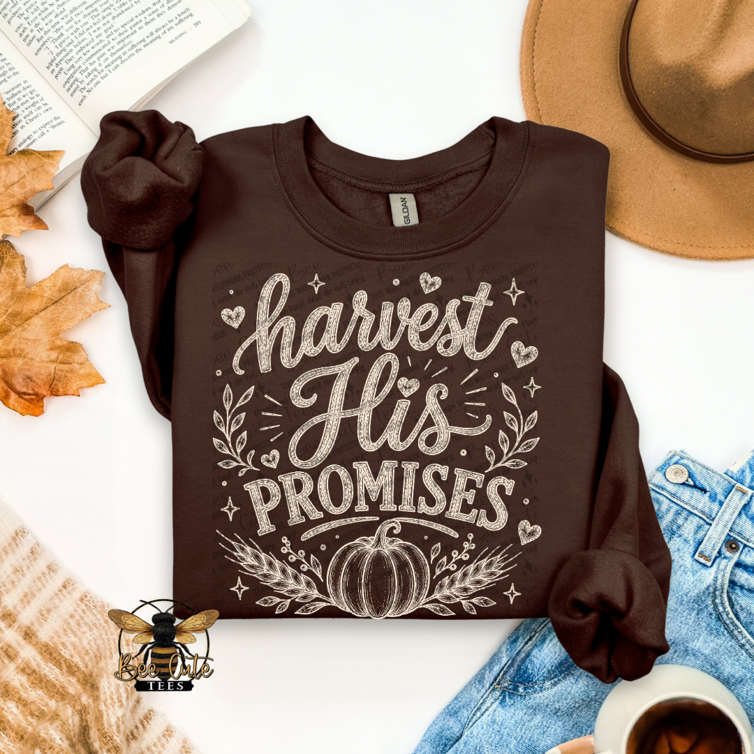 A dark brown long-sleeve t-shirt with a 'harvest His PROMISES' graphic printed in white, styled in a flat-lay arrangement with autumn leaves, a tan hat, blue jeans, a cup of coffee, and a book on a white background.