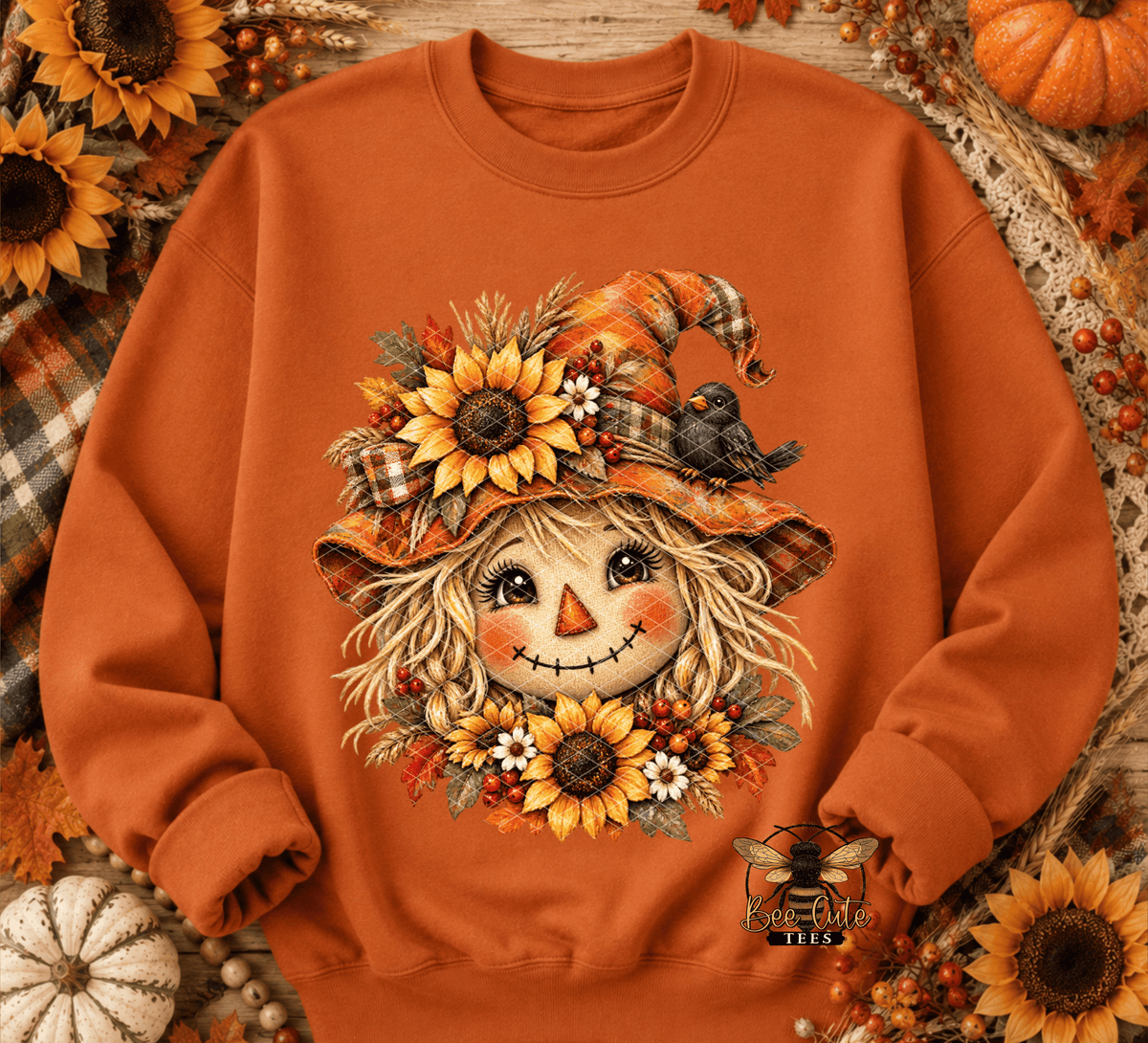 An orange crewneck sweatshirt featuring a detailed autumn scarecrow graphic on the front, surrounded by sunflowers, pumpkins, and a rustic wood background.