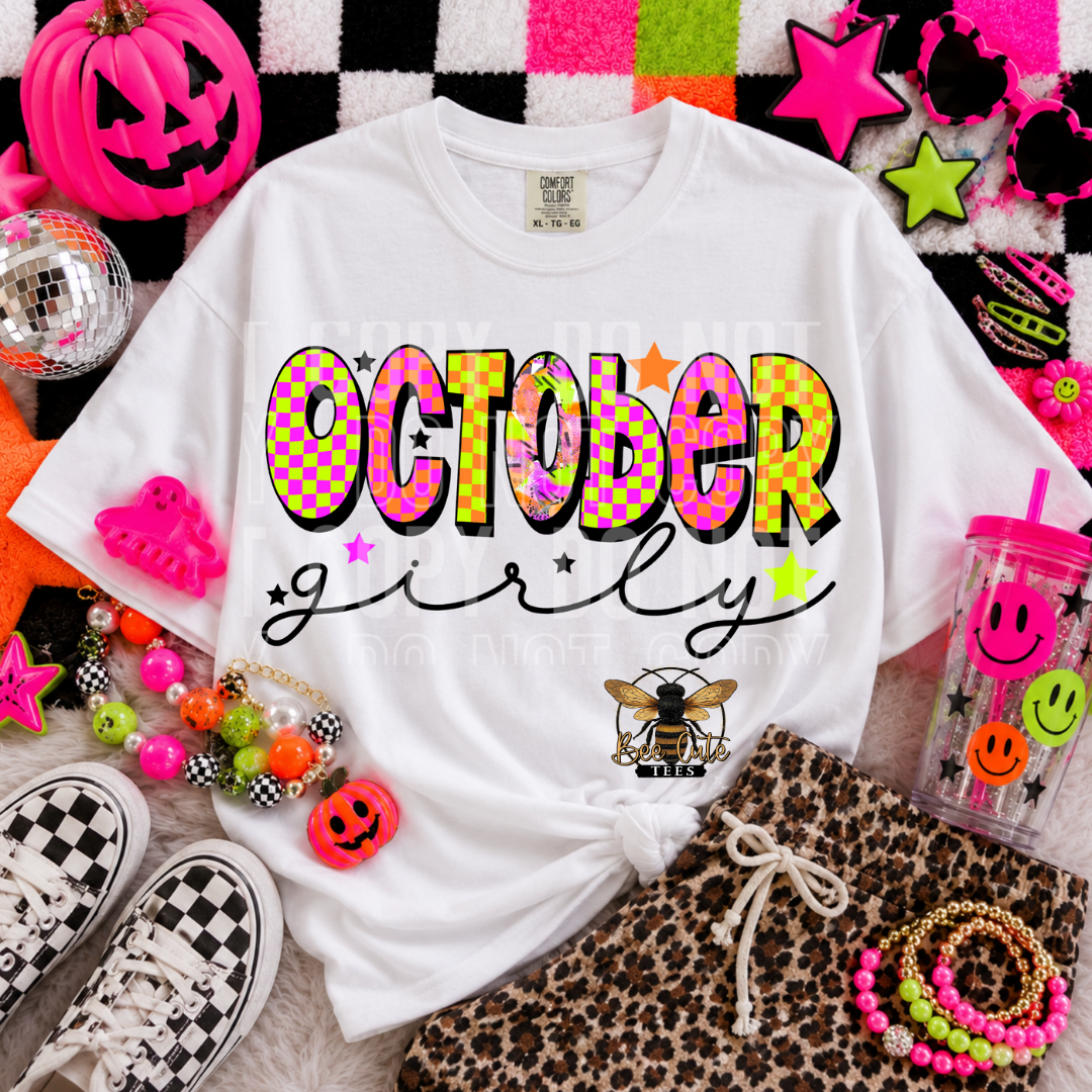 A white graphic T-shirt laid flat in a styled Halloween-themed flat lay, with neon pink and multicolor accessories around it. The shirt reads “OCTOBER girly” in large colorful letters with stars and a small bee graphic near the lower right front.