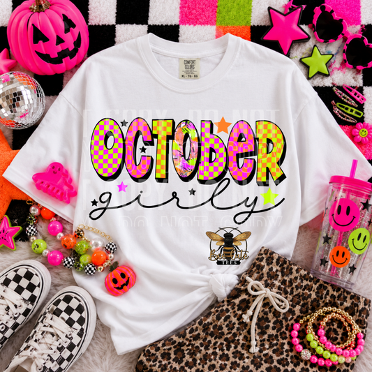 A white graphic T-shirt laid flat in a styled Halloween-themed flat lay, with neon pink and multicolor accessories around it. The shirt reads “OCTOBER girly” in large colorful letters with stars and a small bee graphic near the lower right front.
