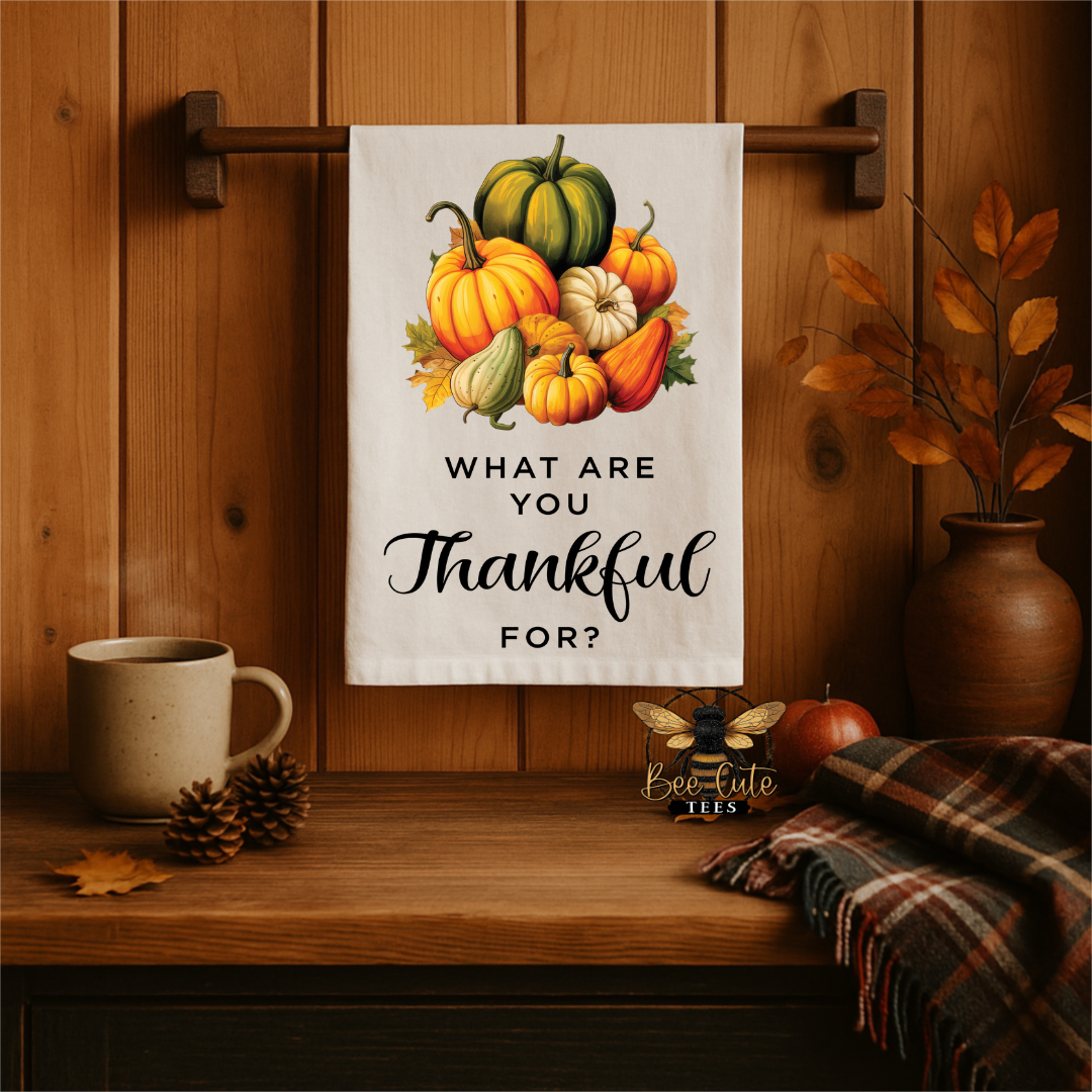 A white kitchen tea towel with a colorful autumn pumpkin illustration and the text 'WHAT ARE YOU Thankful FOR?' hangs on a wooden wall rack in a cozy fall-themed setting with a mug, pinecones, a plaid throw, and a vase of leaves.