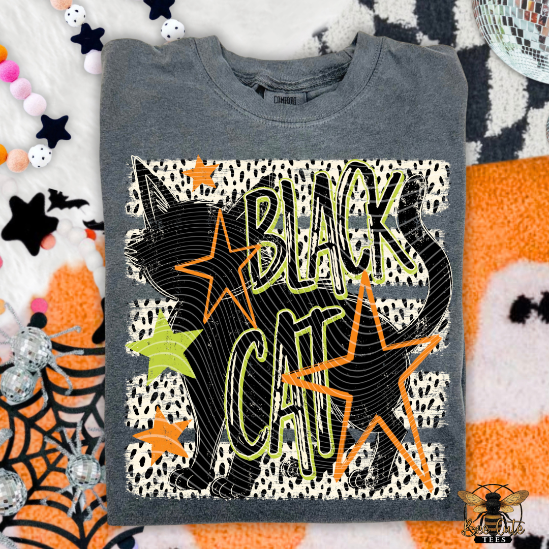A folded dark gray graphic t-shirt featuring a black cat silhouette and the words 'BLACK CAT' in outlined lettering over a white spotted pattern, surrounded by orange and lime green stars. The shirt is styled on a festive Halloween-themed background with spiderwebs, stars, and orange-and-white patterns.