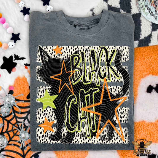 A folded dark gray graphic t-shirt featuring a black cat silhouette and the words 'BLACK CAT' in outlined lettering over a white spotted pattern, surrounded by orange and lime green stars. The shirt is styled on a festive Halloween-themed background with spiderwebs, stars, and orange-and-white patterns.