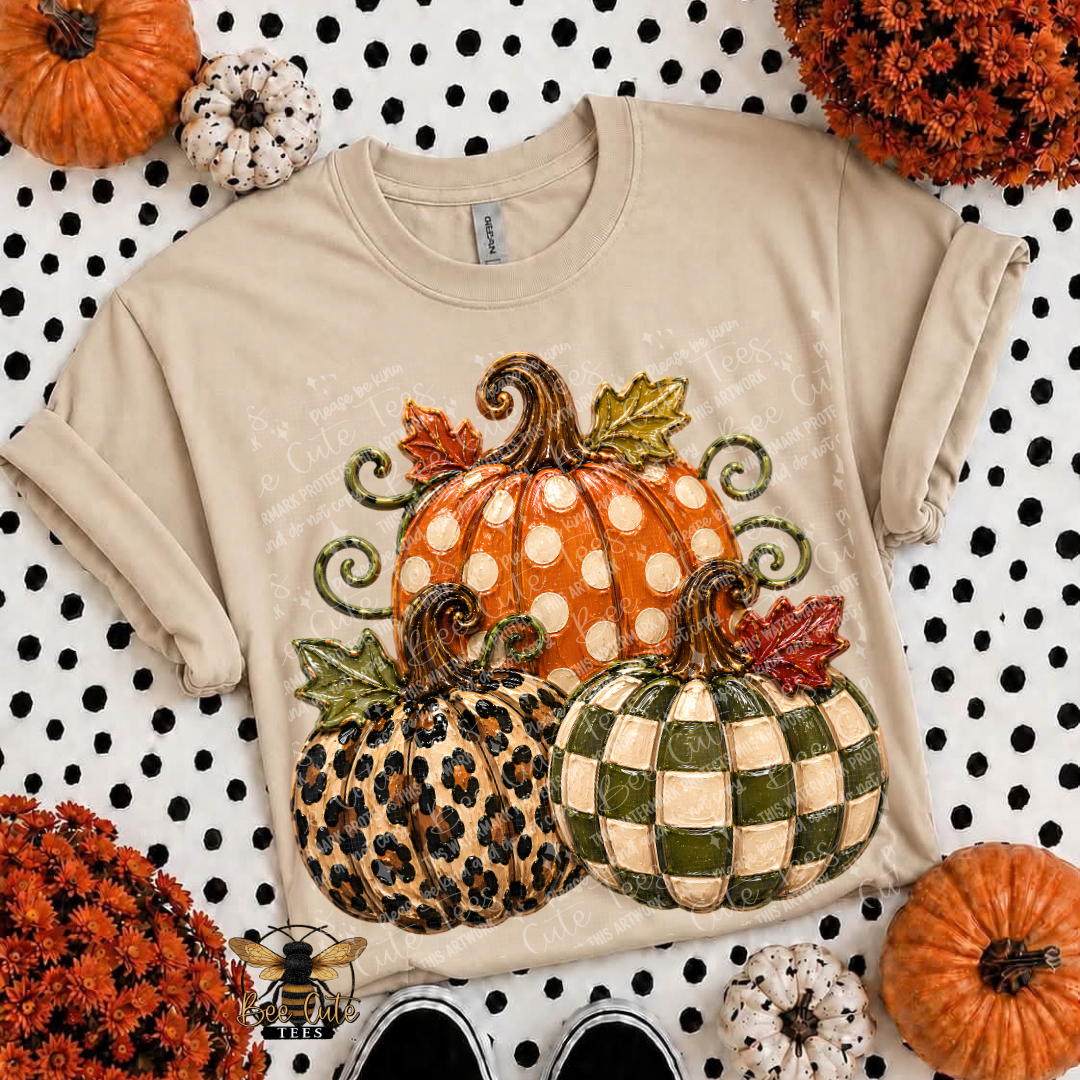 A beige short-sleeve t-shirt with a large autumn pumpkin graphic on the front, styled in a flat-lay with pumpkins, mums, and black-and-white polka dot fabric.