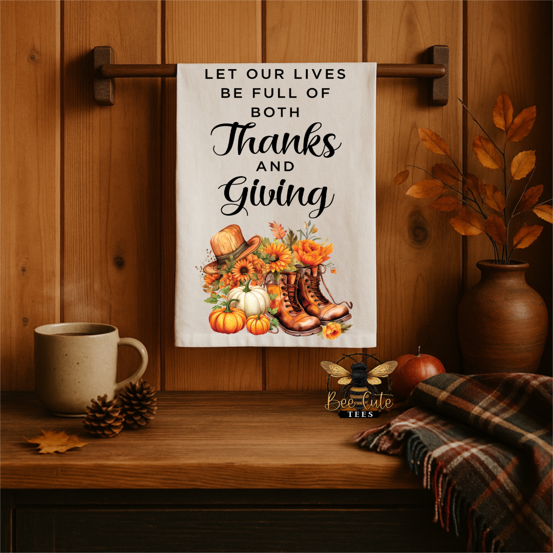 A white kitchen towel with a Thanksgiving-themed print hangs from a wooden rack on a wood-paneled wall. The towel features the text 'LET OUR LIVES BE FULL OF BOTH Thanks AND Giving' above an illustration of autumn elements including pumpkins, sunflowers, a cowboy hat, boots, and leaves.