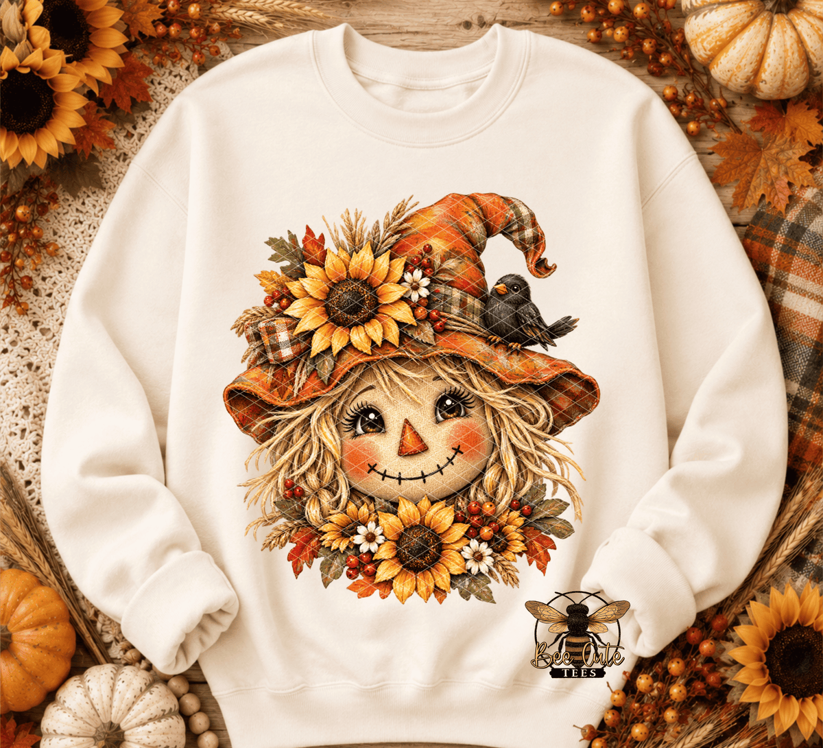 An off-white crewneck sweatshirt featuring a detailed autumn scarecrow graphic on the front, surrounded by sunflowers, leaves, and a small bird, with a bee logo near the lower right sleeve. The sweatshirt is styled in a flat-lay with seasonal props like pumpkins, sunflowers, and a plaid blanket.