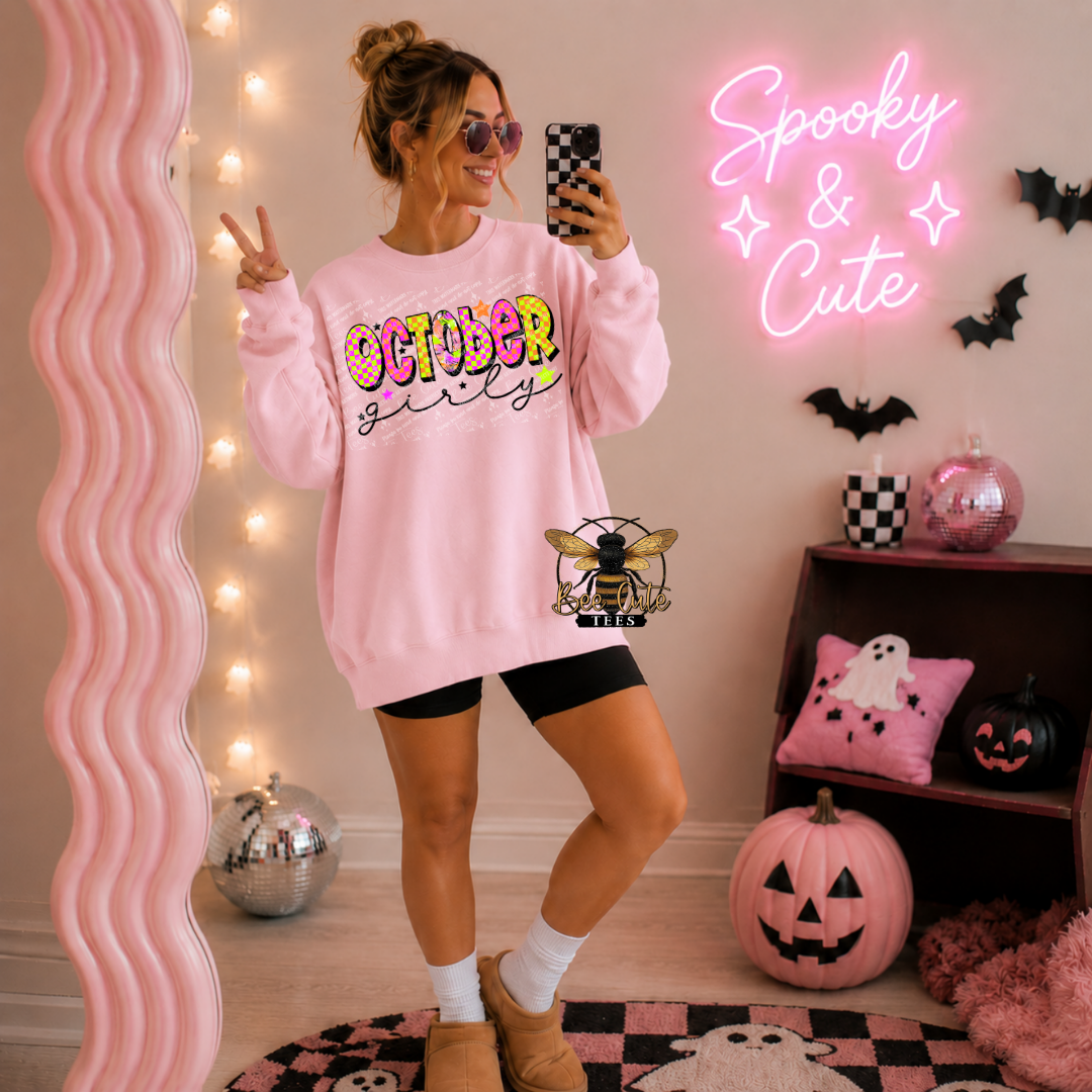 A woman takes a mirror selfie wearing a pink crewneck sweatshirt with 'OCTOBER Girly' written in colorful letters and a bee graphic on the front, paired with black bike shorts. The room is decorated for Halloween with a pink neon sign, pumpkins, and string lights.
