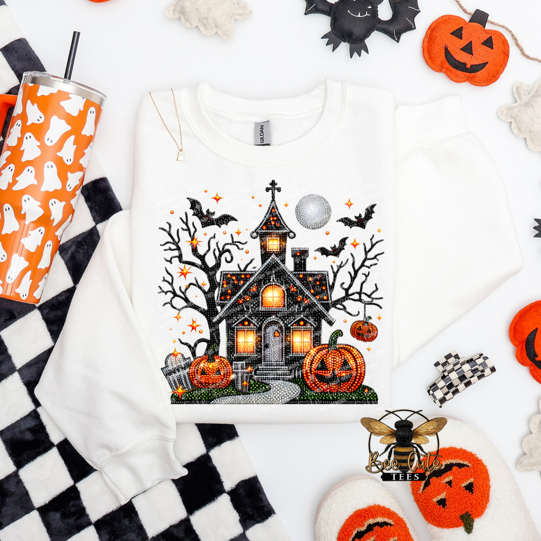 A white long-sleeve sweatshirt laid flat on a white background, decorated with a large Halloween graphic featuring a haunted house, pumpkins, bats, stars, and a full moon. The sweatshirt is styled in a flat lay with seasonal props including a black-and-white checkered cloth, an orange cup with ghost patterns, black bat cutouts, and orange pumpkin decorations.