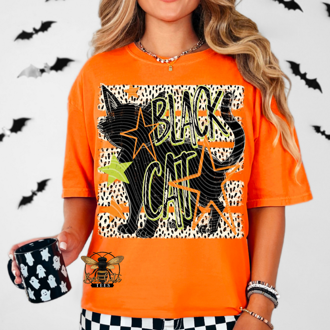 A woman wearing a bright orange graphic t-shirt with a black cat, leopard print, and the words 'BLACK CAT' in green and yellow. She is holding a black mug with white ghost patterns.