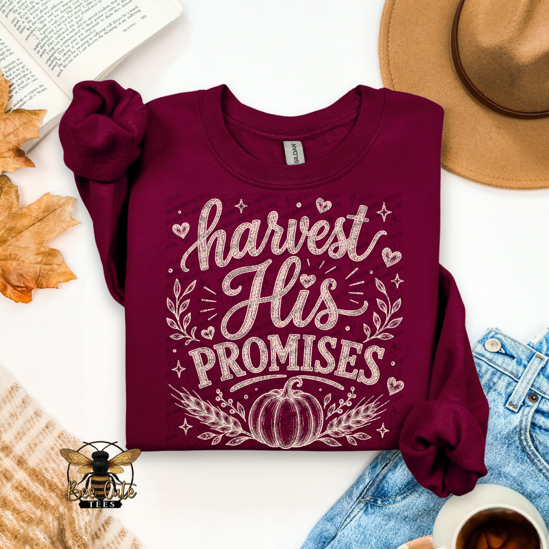 A folded burgundy long-sleeve t-shirt with a white graphic reading 'harvest His PROMISES' with a pumpkin and decorative leaves, styled in a flat lay with a hat, jeans, leaves, a book, and a cup on a white background.