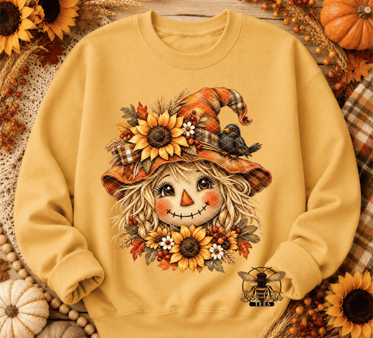 A mustard yellow crewneck sweatshirt featuring a detailed autumn scarecrow graphic on the front. The scarecrow has a patchwork orange hat decorated with sunflowers, a small black bird, and autumn leaves.