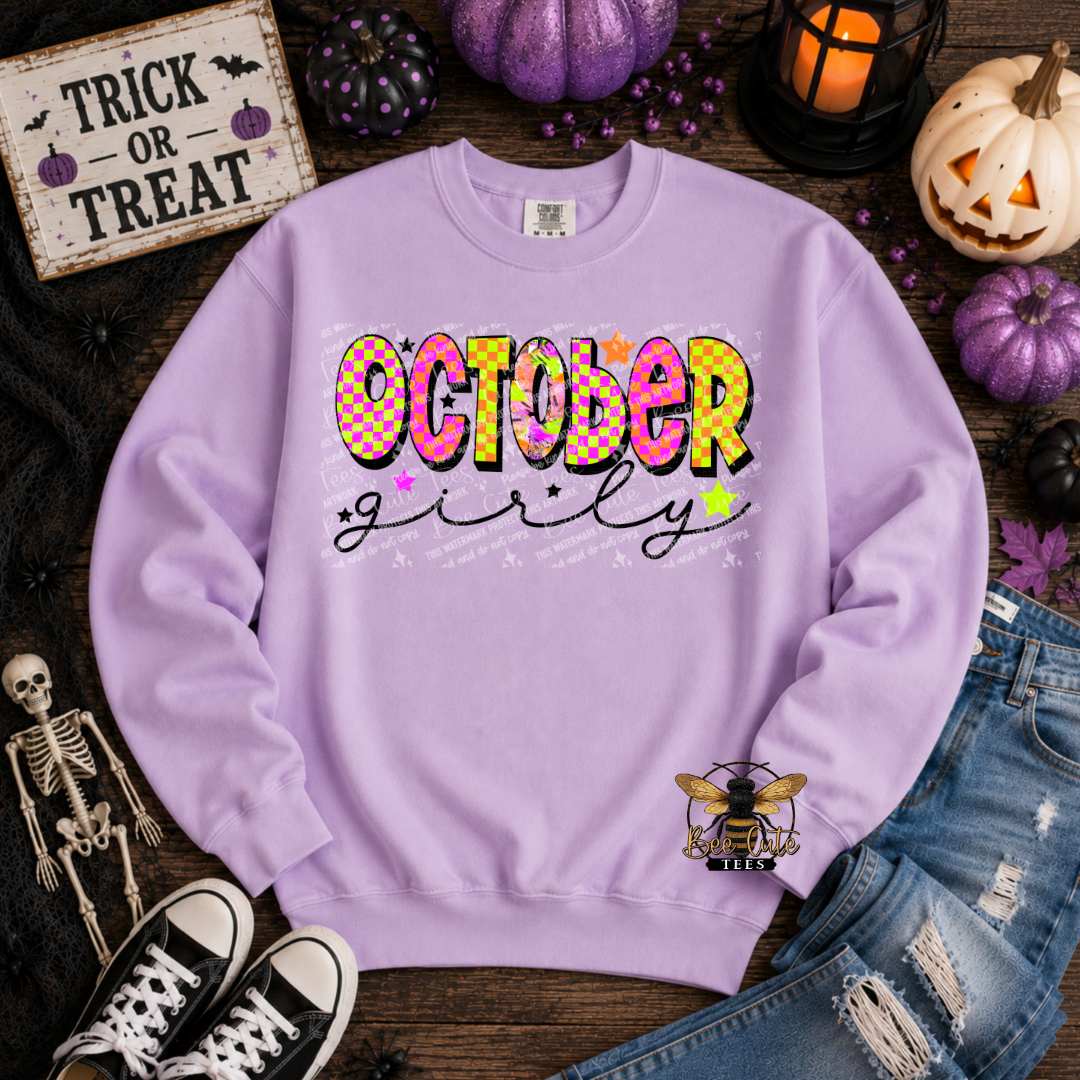A lavender crewneck sweatshirt with a colorful 'October girly' graphic on the chest, styled in a flat lay with Halloween props including pumpkins, a skeleton, jeans, and sneakers.
