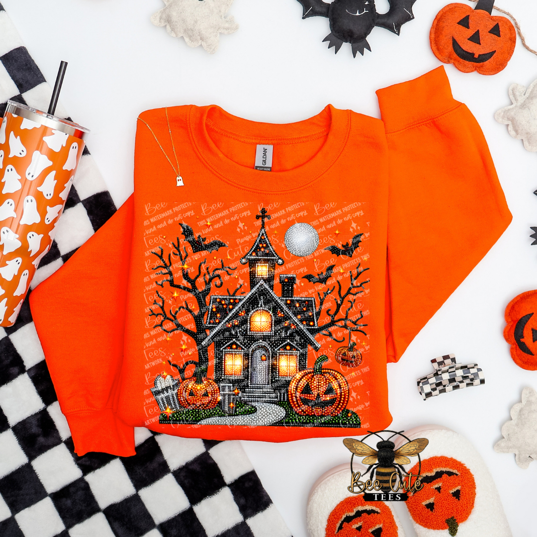 An orange crewneck sweatshirt with a detailed Halloween haunted house graphic, styled in a flat lay with seasonal props like jack-o'-lanterns, a checkered blanket, and a ghost-patterned tumbler.