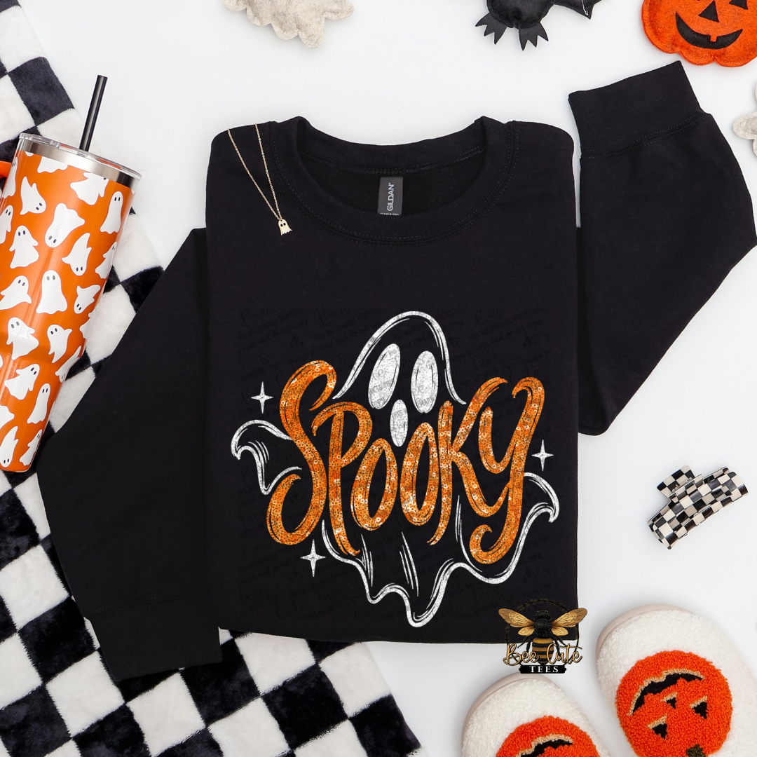 A black crewneck sweatshirt with a glittery orange 'Spooky' graphic and a white ghost outline, styled in a flat lay with Halloween props including a pumpkin cup, checkered blanket, and orange socks.