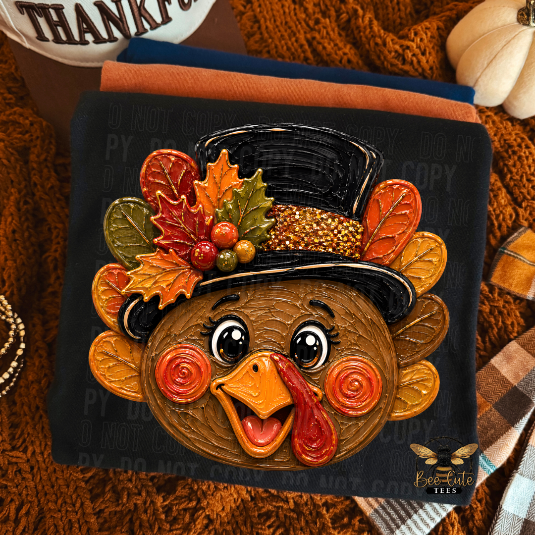 A folded black t-shirt featuring a stylized cartoon turkey face wearing a black top hat decorated with autumn leaves and berries, shown on a dark fabric background with a cozy fall-themed styled flat lay.