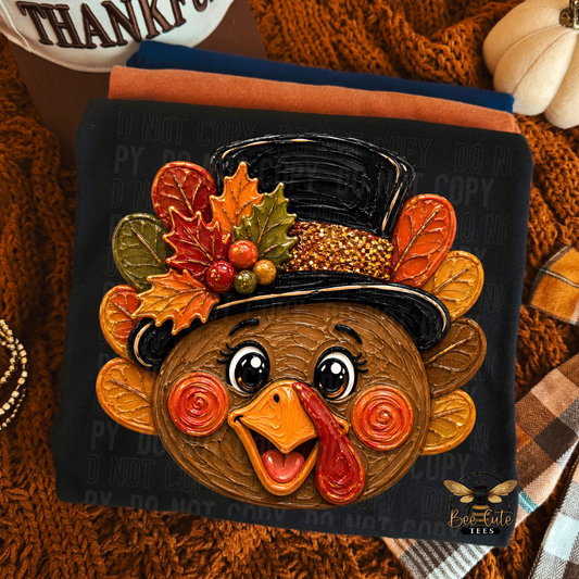 A folded black t-shirt featuring a stylized cartoon turkey face wearing a black top hat decorated with autumn leaves and berries, shown on a dark fabric background with a cozy fall-themed styled flat lay.