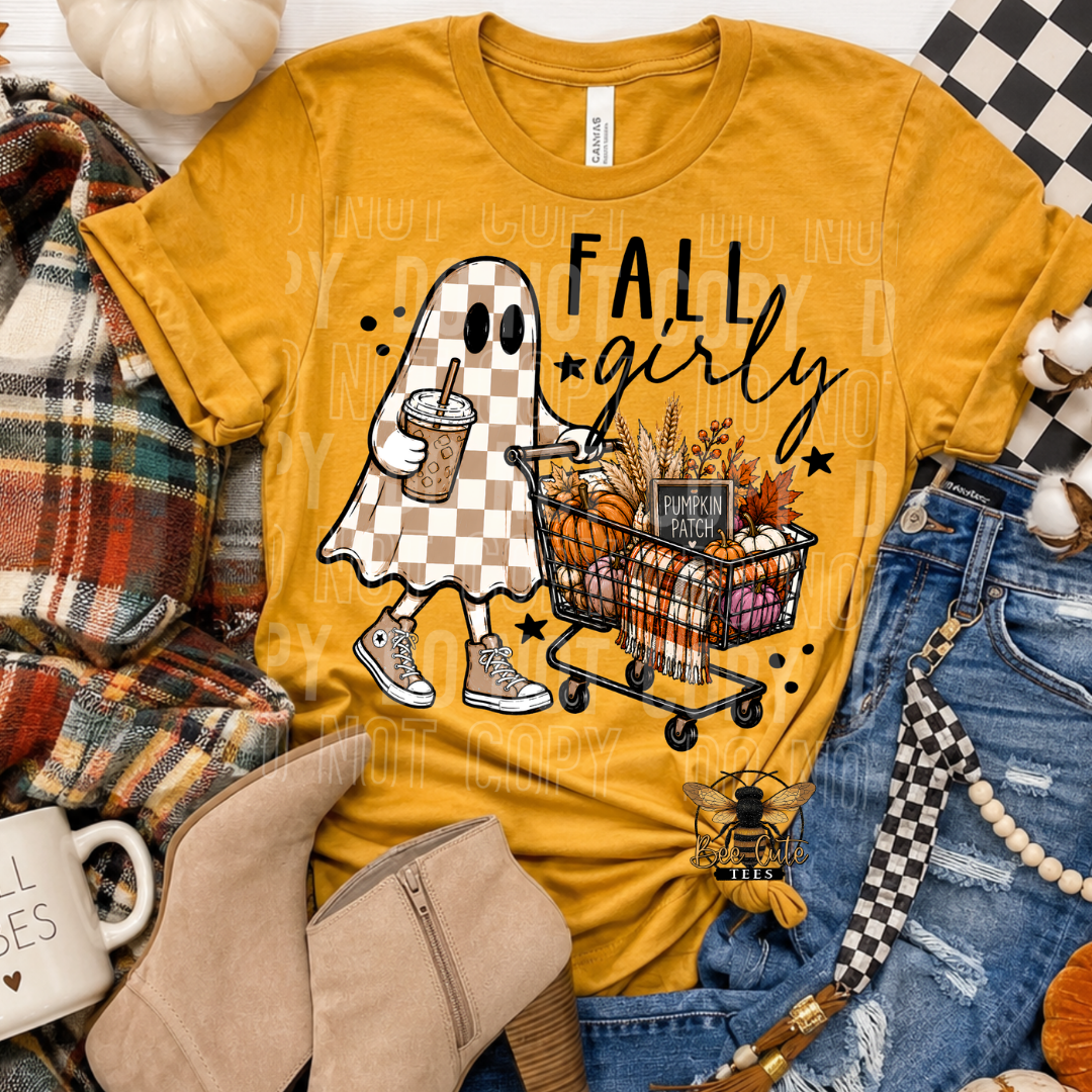 A mustard yellow graphic T-shirt laid flat with autumn styling props. The shirt features a ghost character in a checkerboard pattern pushing a shopping cart filled with fall elements, with the words “FALL girly” printed on the front.