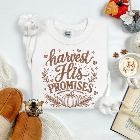 A white long-sleeve t-shirt with a brown graphic reading 'harvest His PROMISES' and a pumpkin design, styled in an autumn flat lay with slippers, a mug of coffee, an open book, and fall leaves.