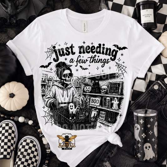 A white graphic t-shirt with a black and white skeleton in a shopping cart scene, featuring the text 'just needing a few things' and a small bee logo at the bottom. The t-shirt is styled in a flat-lay arrangement with Halloween-themed props including a pumpkin, checkered shoes, a tumbler, and a candle.