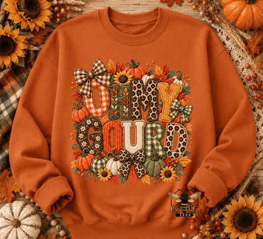 An orange crewneck sweatshirt featuring a 'Oh My Gourd' autumn graphic with pumpkins, sunflowers, and patterned letters, styled in a flat lay with pumpkins and sunflowers.
