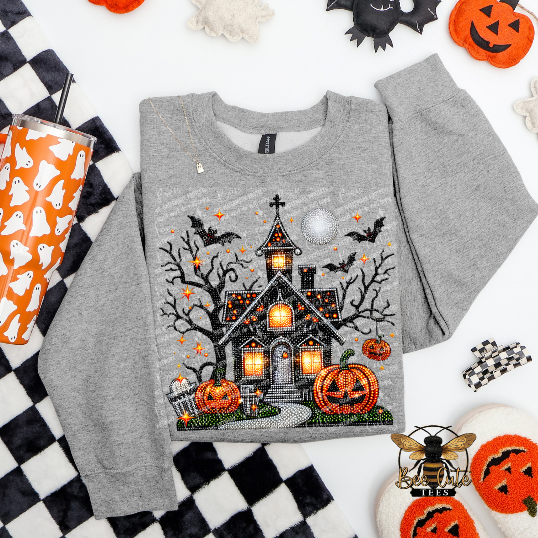 A heather gray long-sleeve sweatshirt with a detailed Halloween graphic of a haunted house, pumpkins, bats, and a full moon, styled in a flat lay with seasonal props.