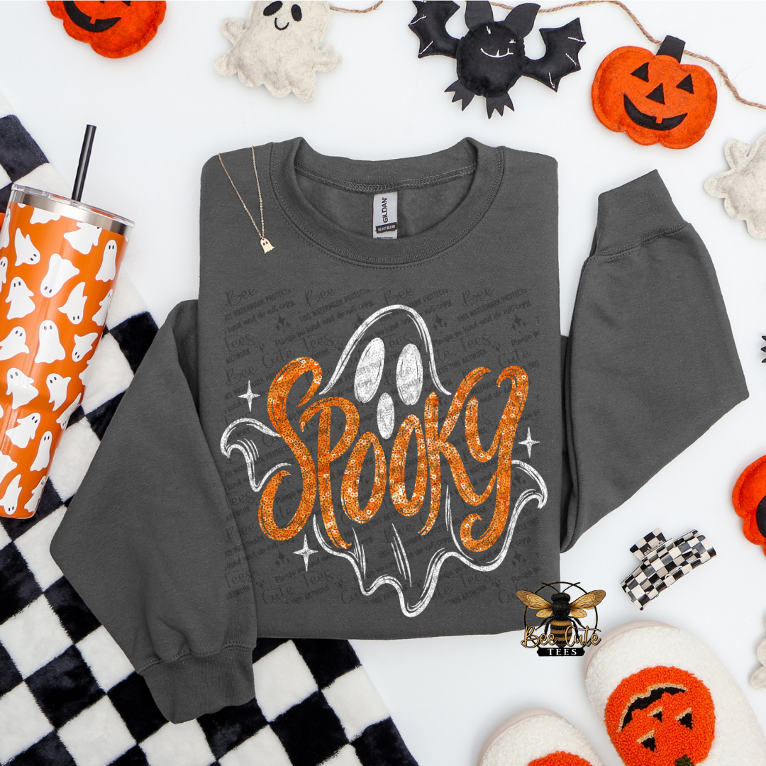 A dark gray crewneck sweatshirt with a 'Spooky' ghost graphic, styled in a flat lay with Halloween props including jack-o'-lanterns, a tumbler, and a checkered blanket.