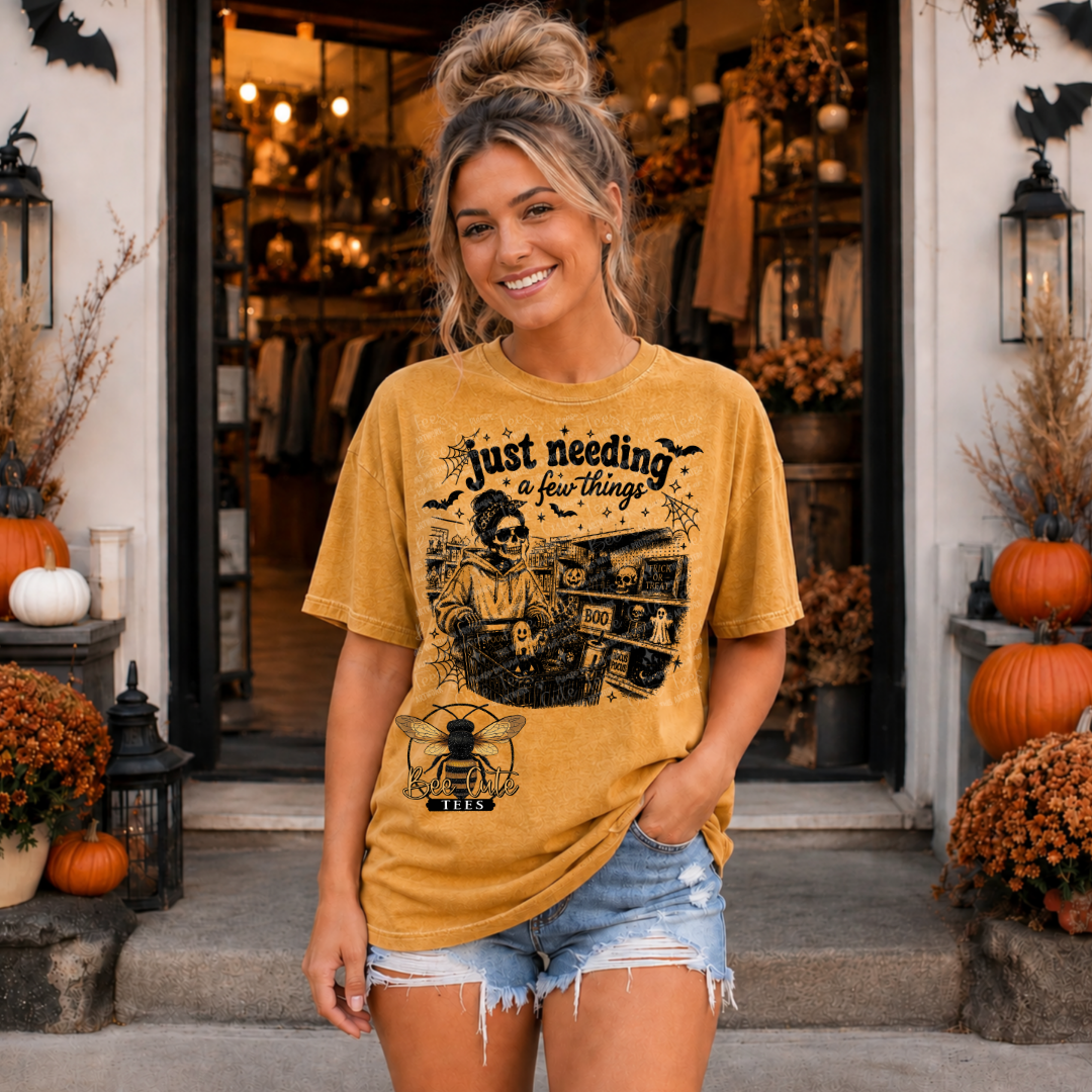 A woman stands in front of a Halloween-themed storefront doorway wearing a mustard yellow graphic t-shirt with a black print that reads 'Just needing a few things' above a spooky scene with bats and a bee. She is also wearing denim shorts.