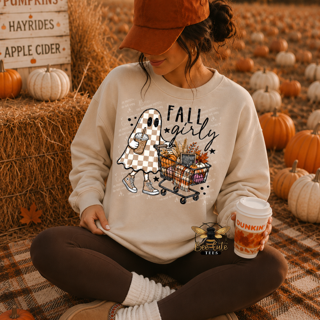 A woman sitting cross-legged in an autumn pumpkin yard wears a cream sweatshirt with a 'Fall Girty' cartoon ghost graphic. She is wearing a rust-colored cap, dark brown leggings, and holding a Dunkin' coffee cup.