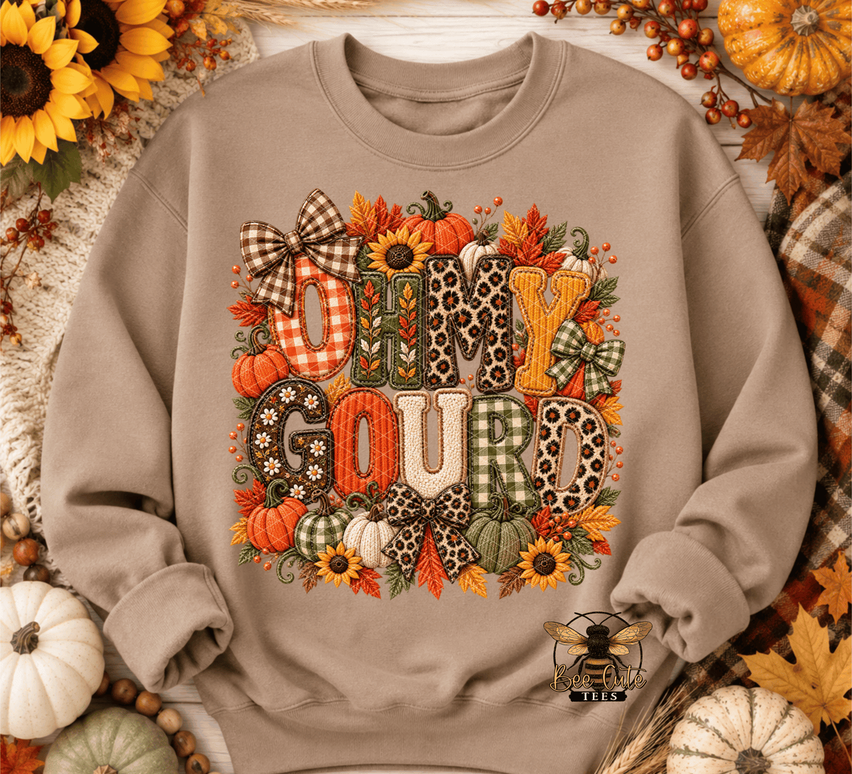 A taupe crewneck sweatshirt featuring a 'Oh My Gourd' autumn graphic with patterned letters, pumpkins, sunflowers, and bows, styled in a flat lay with fall decorations.
