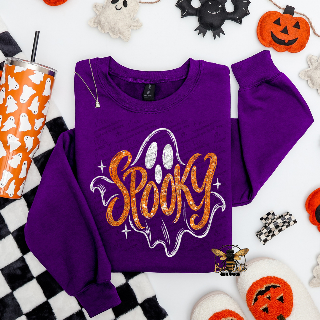 A purple crewneck sweatshirt with a glittery orange and white 'Spooky' graphic, styled in a flat lay with Halloween-themed props including a pumpkin, ghost decorations, a tumbler, and slippers on a white background.