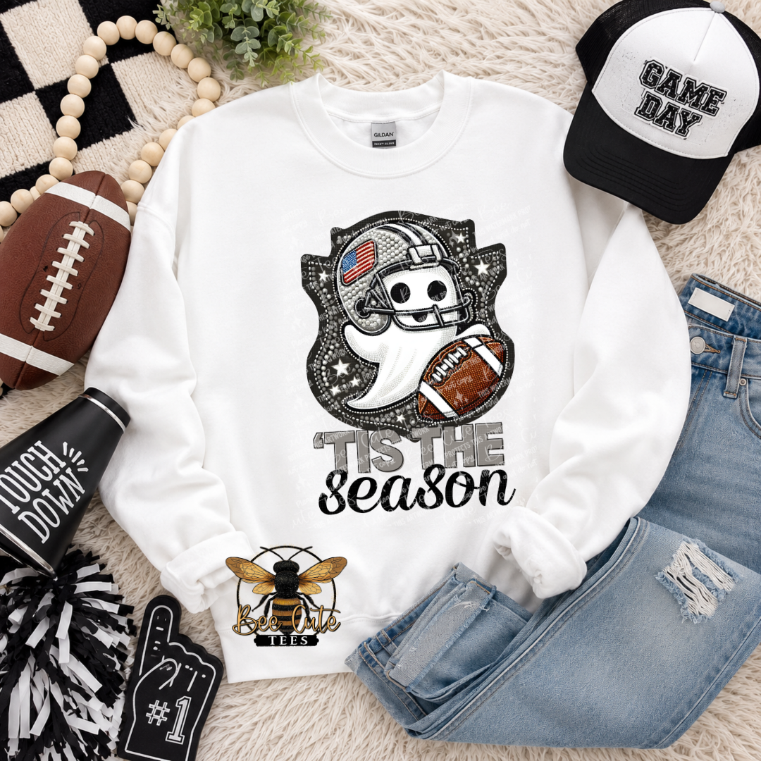 A white long-sleeve sweatshirt laid flat on a cream rug with seasonal props. The sweatshirt has a black and white football-themed ghost graphic with the text 'TIS THE season.
