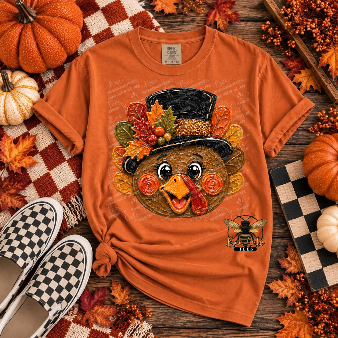 An orange short-sleeve graphic T-shirt laid flat on a rustic wood surface with autumn props. The shirt features a large turkey face wearing a black top hat with colorful fall leaves and berries, plus a small bee graphic near the lower right front.