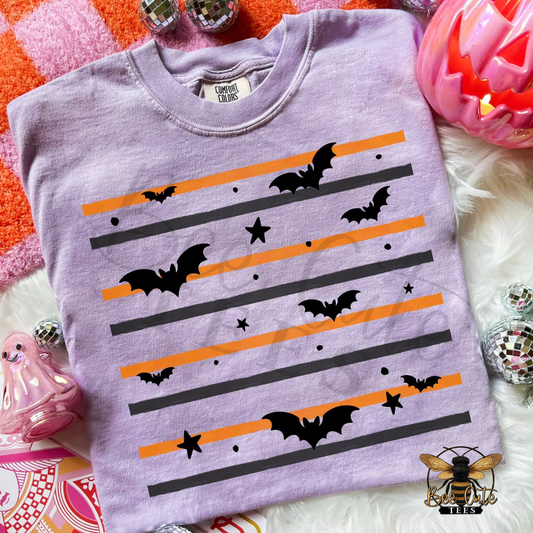 A lavender crewneck t-shirt with a Halloween graphic print of black bats, stars, and diagonal orange and black stripes, styled on a white faux fur surface with festive props.