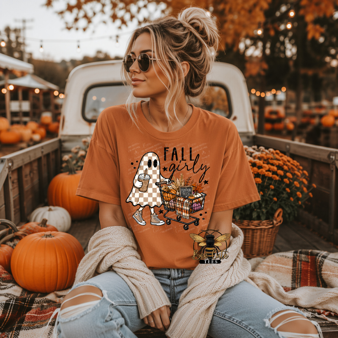 A woman wearing a burnt orange graphic t-shirt sits in the bed of a pickup truck surrounded by pumpkins and fall decorations. The t-shirt features a cartoon ghost shopping for Halloween with the text 'FALL girly'.