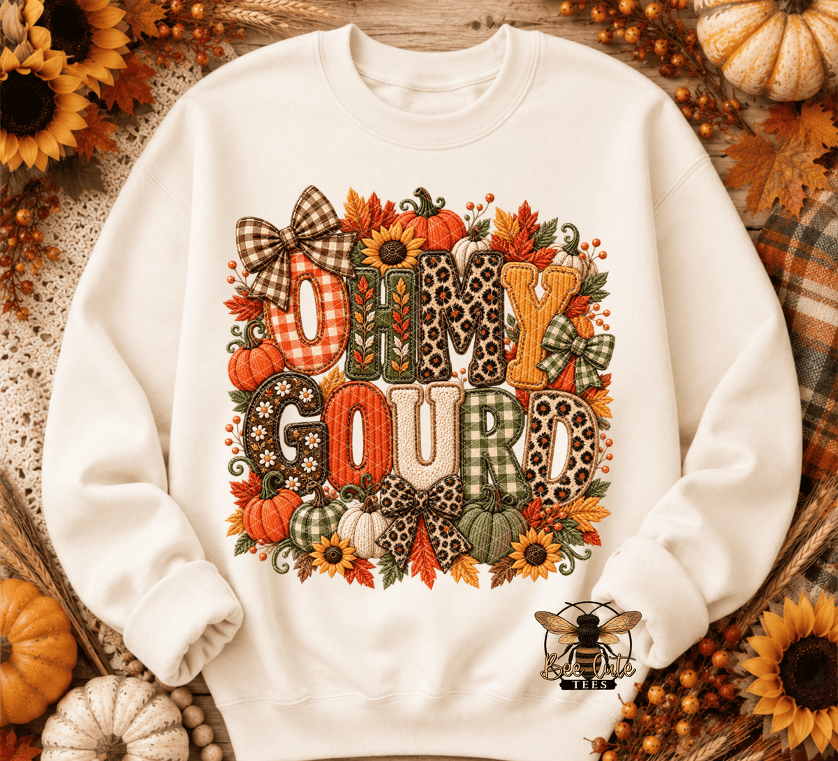 An off-white crewneck sweatshirt featuring a large, colorful 'Oh My Gourd' graphic with autumn-themed patterns like leopard print, gingham, and plaid, surrounded by pumpkins, sunflowers, and leaves. The sweatshirt is displayed in a flat-lay arrangement with seasonal props including sunflowers, pumpkins, and a plaid blanket.
