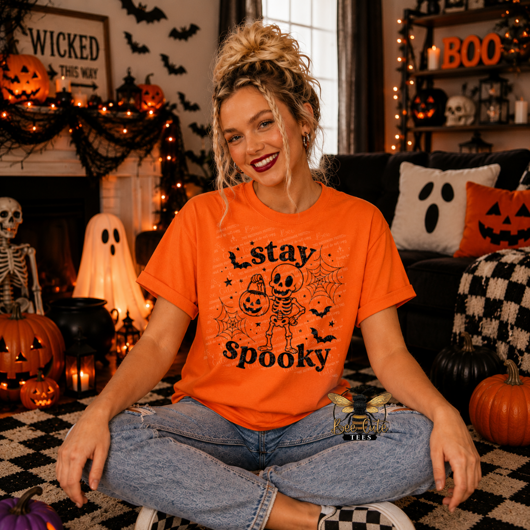 A woman sitting cross-legged in a cozy, decorated Halloween living room, wearing a bright orange graphic t-shirt with a black skeleton and jack-o'-lantern design that reads 'stay spooky'.
