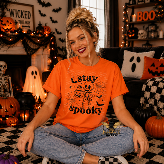 A woman sitting cross-legged in a cozy, decorated Halloween living room, wearing a bright orange graphic t-shirt with a black skeleton and jack-o'-lantern design that reads 'stay spooky'.