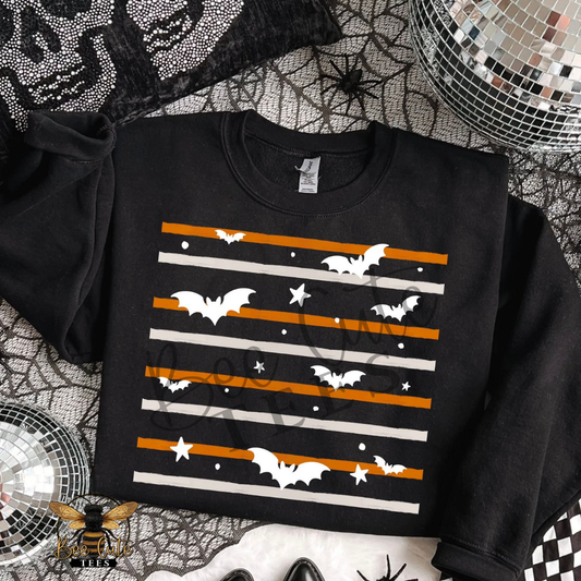 A black long-sleeve sweatshirt laid flat on a Halloween-themed background. The sweatshirt features a graphic print of white bats, white stars, and horizontal orange and white stripes.