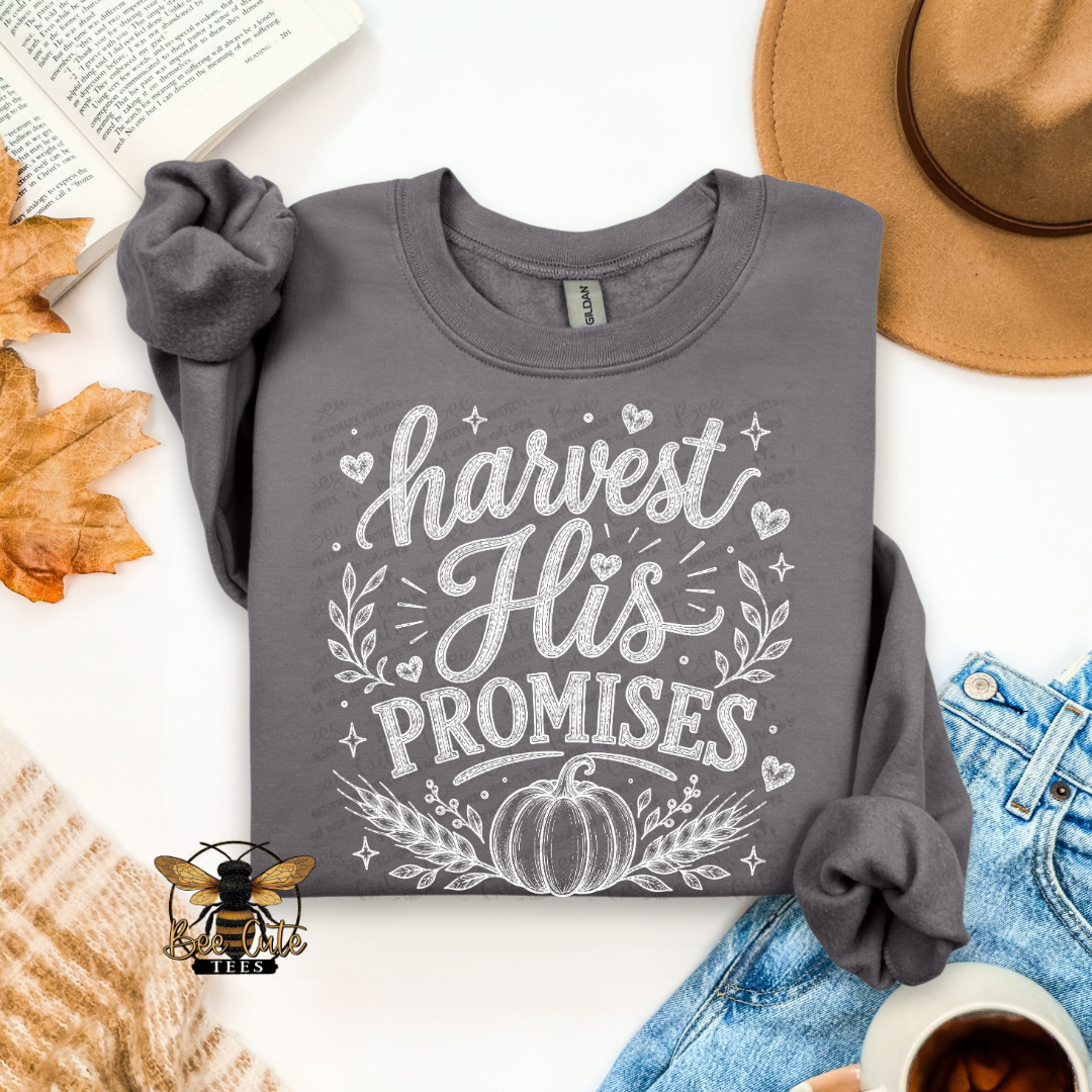 A folded dark gray long-sleeve t-shirt with rolled cuffs, featuring a white graphic print that reads 'harvest His PROMISES' with a pumpkin and leaf design. The shirt is styled in a flat-lay arrangement with autumn leaves, an open book, a tan hat, blue jeans, a cup of coffee, and a knit sweater on a white background.