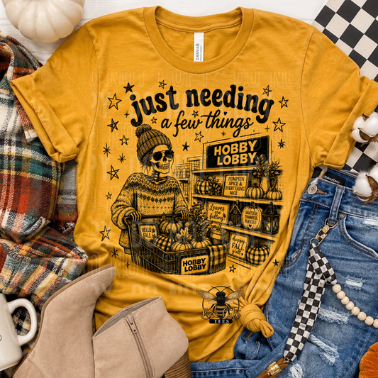 A mustard yellow graphic t-shirt featuring a Fall/Halloween-themed illustration of a skeleton shopper at a store counter with the text 'just needing a few things'. The shirt is styled in a flat-lay arrangement with denim shorts, a plaid scarf, beige boots, and a white pumpkin.