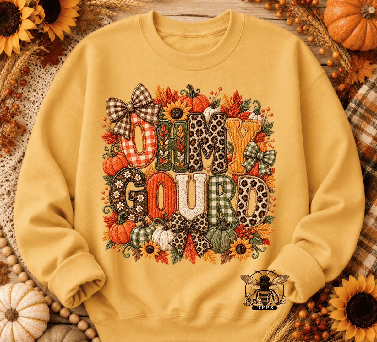 A yellow crewneck sweatshirt with a large 'Oh My Gourd' autumn graphic, styled in a flat lay with fall decorations like sunflowers, pumpkins, and plaid fabrics.