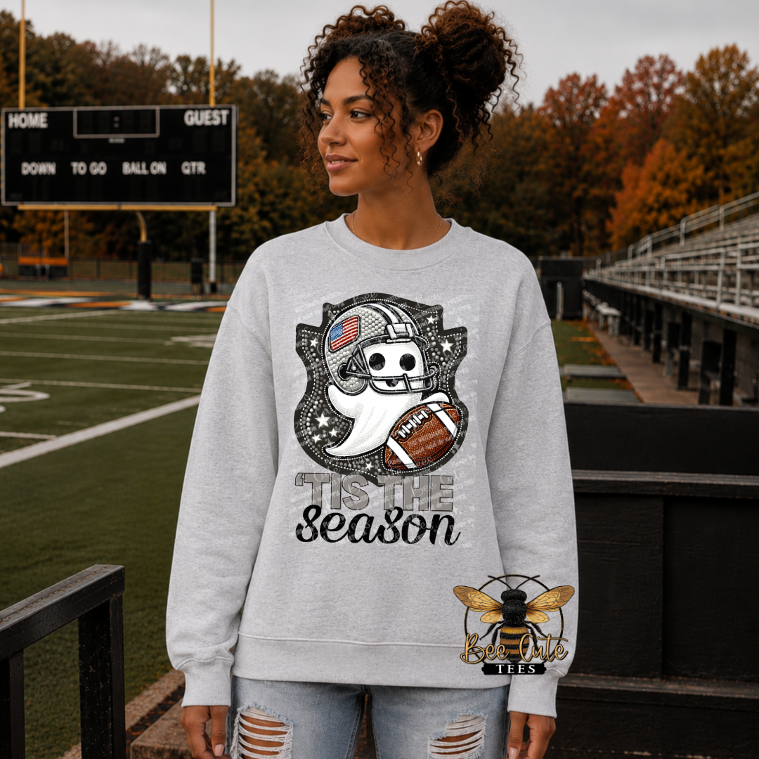 A woman stands on a football field wearing a light gray crewneck sweatshirt with a Halloween-themed graphic that reads 'Tis the Season' and shows a skull in a football helmet, a ghost, and a football. The background shows a blurred sports field and trees.