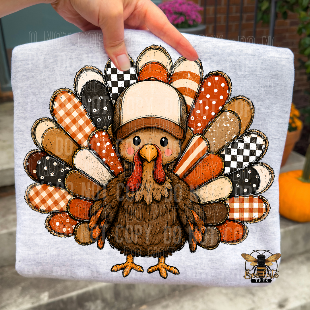 A hand holds up a light heather gray T-shirt featuring a large embroidered-style turkey graphic with patterned feathers in autumn colors. The background is an outdoor patio setting with a pumpkin and brick wall visible.