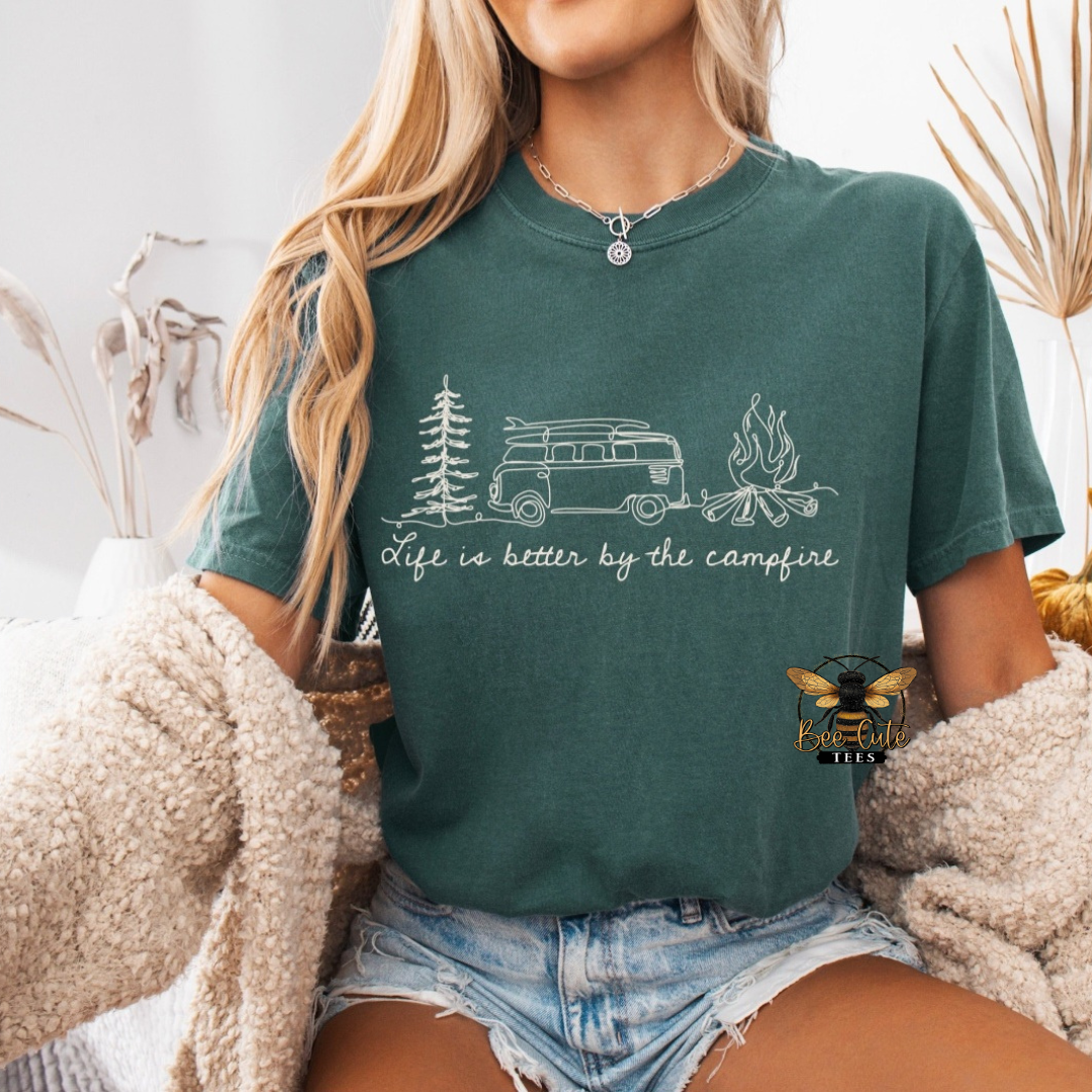 A woman wearing a dark green graphic t-shirt with a white line art design of a van, trees, and campfire, with the text 'life is better by the campfire'. She is wearing denim shorts and a fuzzy cream knit cardigan, with a  brand logo watermark in the bottom right corner.